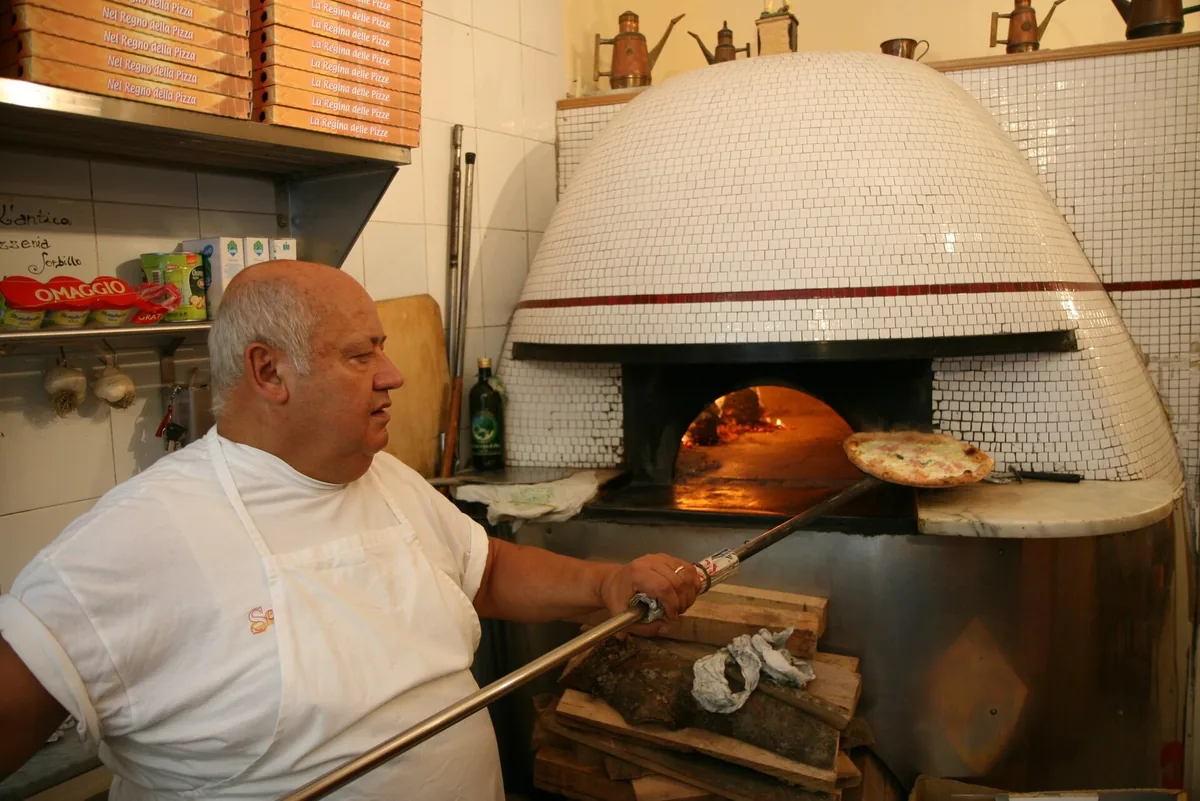 A pizzaiolo sliding a pizza into a tiled wood-fired oven in a Naples pizzeria