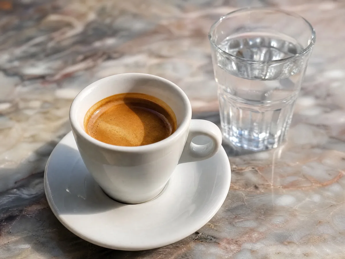 Ein Espresso mit dicker Crema in einer weißen Porzellantasse, daneben ein Glas Wasser