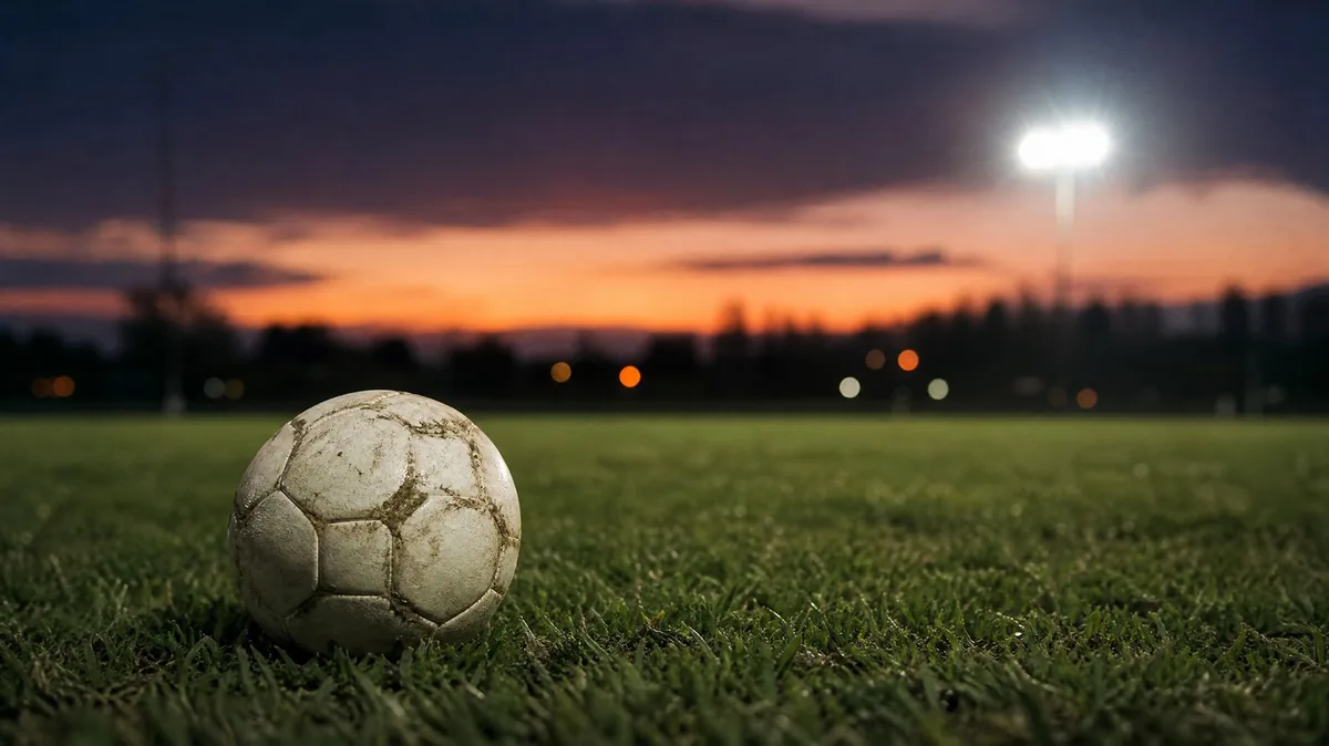 A worn football on the grass of an empty pitch at dusk under floodlights