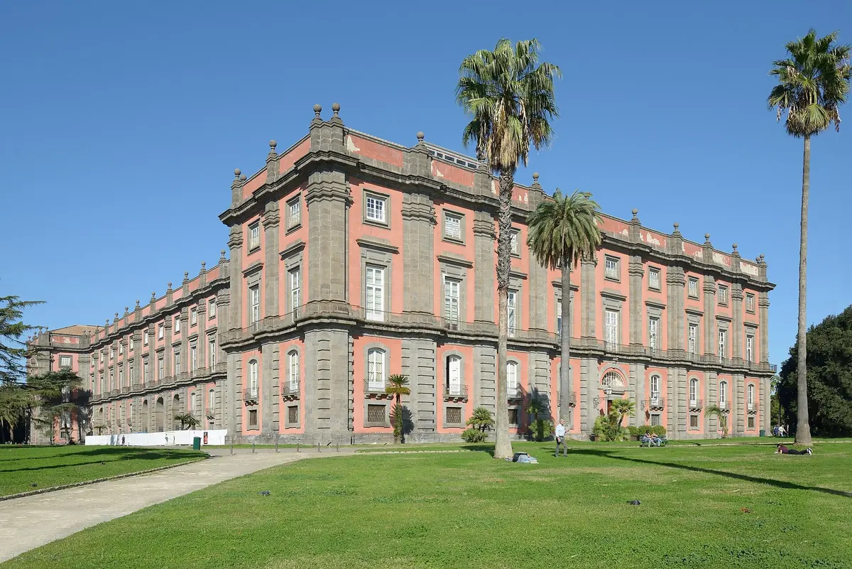 Le palais rouge et gris de la Reggia di Capodimonte derrière une pelouse et de hauts palmiers
