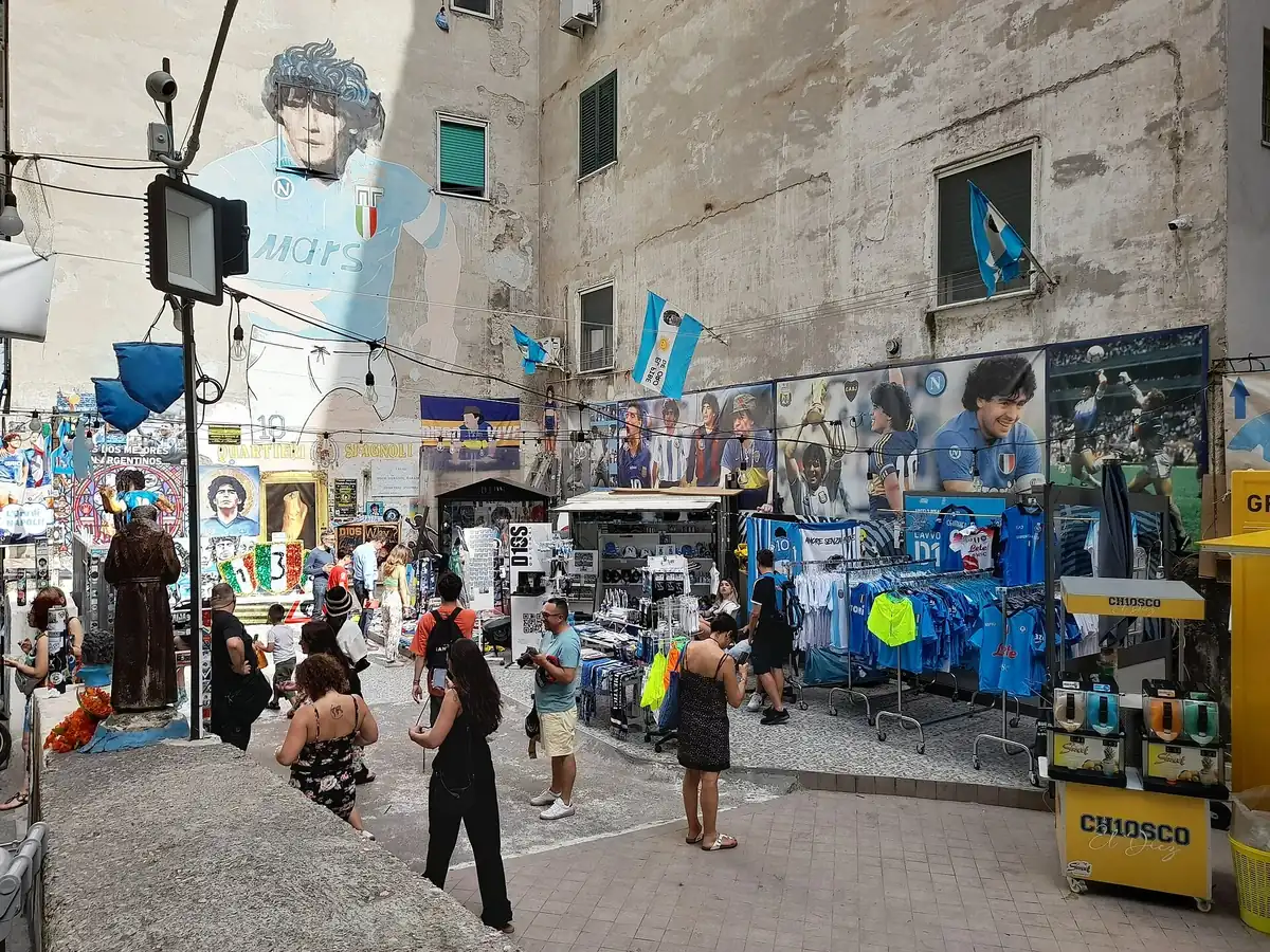 Visiteurs et étals de souvenirs napolitains sous la fresque de Maradona au Largo Maradona